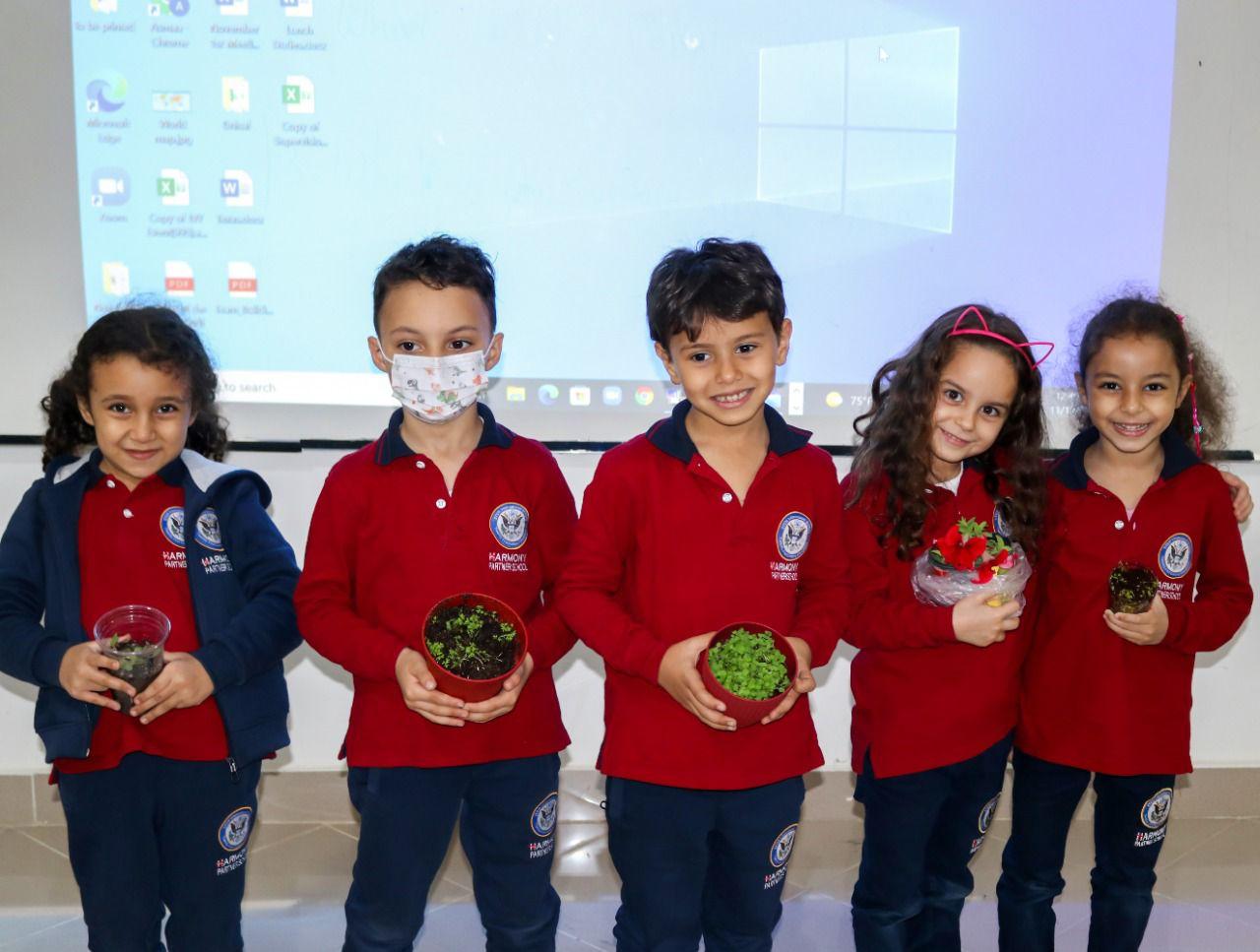 Four students from IVY STEM International School participating in a classroom activity with computers and plants, wearing matching red uniforms and holding small plants.